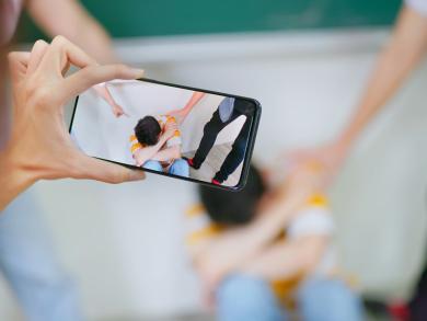 Mobbingsituation wird mit Smartphone gefilmt, Kind sitzt bedrängt am Boden.