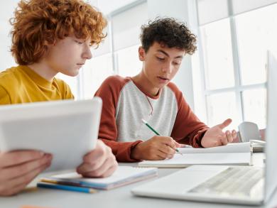 Zwei Jugendliche lernen gemeinsam mit Tablet, Laptop und Heft am Tisch.